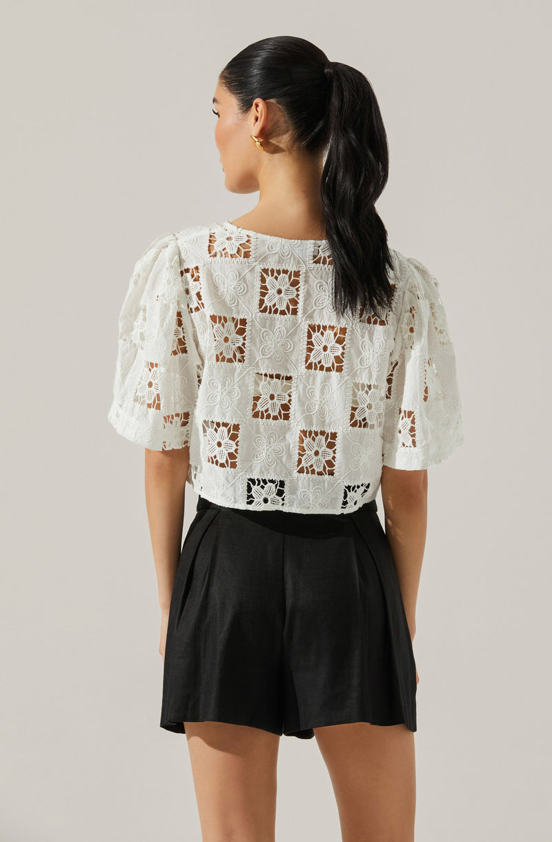 A woman is wearing a white eyelet lace top with puffed short sleeves and a square pattern design, paired with high-waisted black shorts.