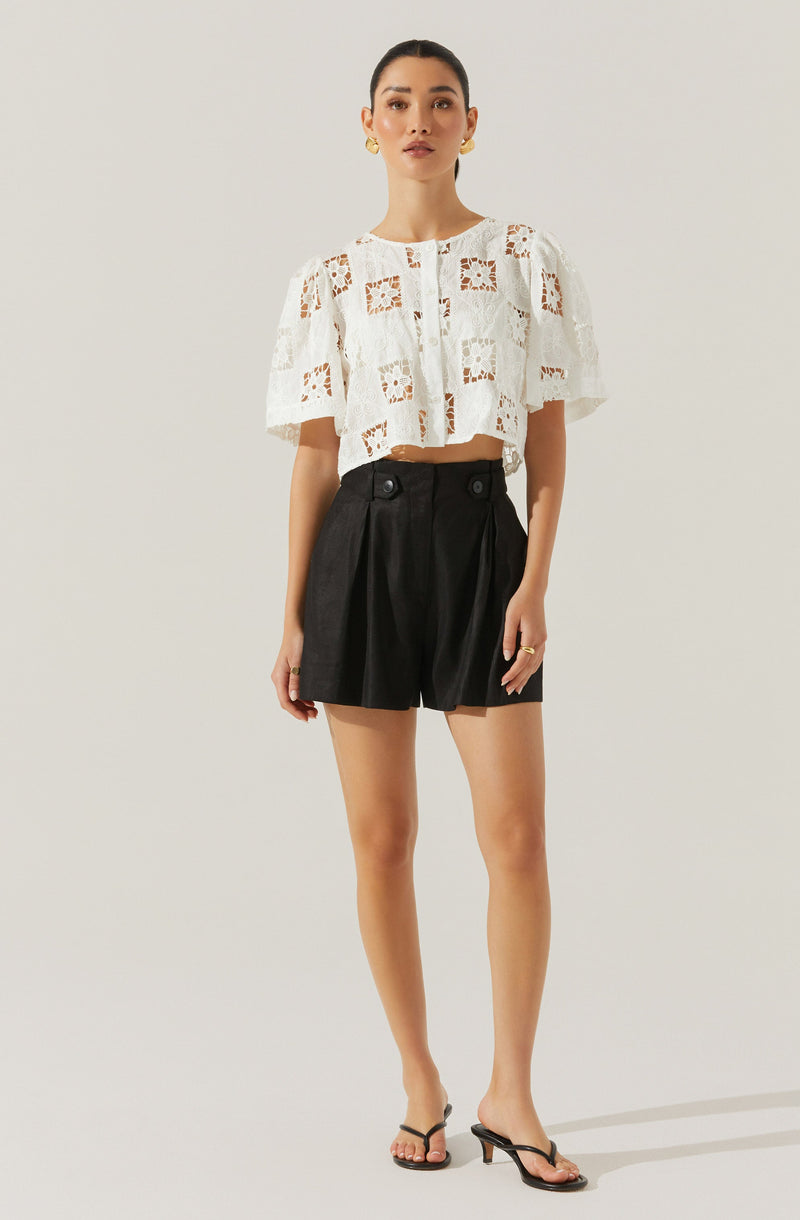 A woman is wearing a white, short-sleeved, lace crop top with a floral pattern and button detailing down the front. She pairs it with high-waisted black shorts that have pleats, wide buttons at the waistband, and a slightly loose fit. She completes the outfit with black strappy heels featuring a small heel.
