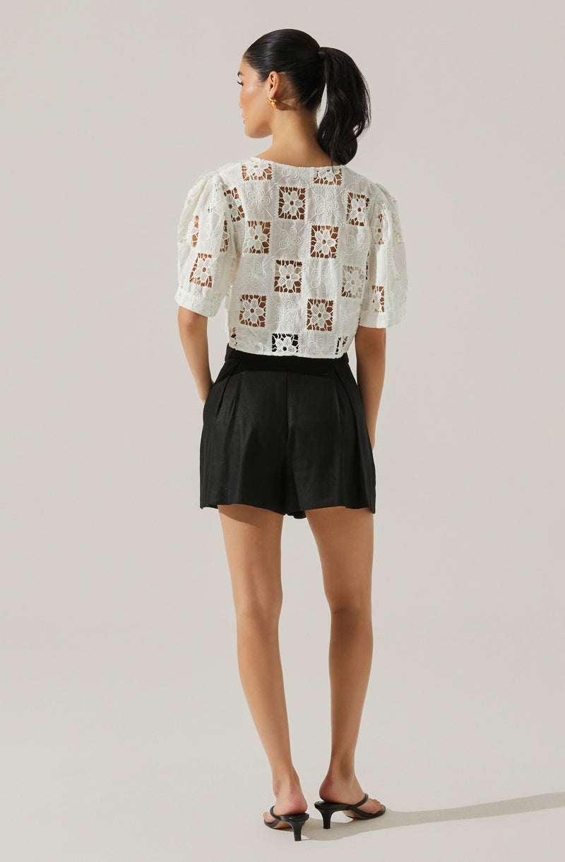 A woman is wearing a white, short-sleeved blouse with a square, cut-out pattern and floral lace design. She has paired it with high-waisted black shorts and black heeled sandals. Her hair is styled in a high ponytail.