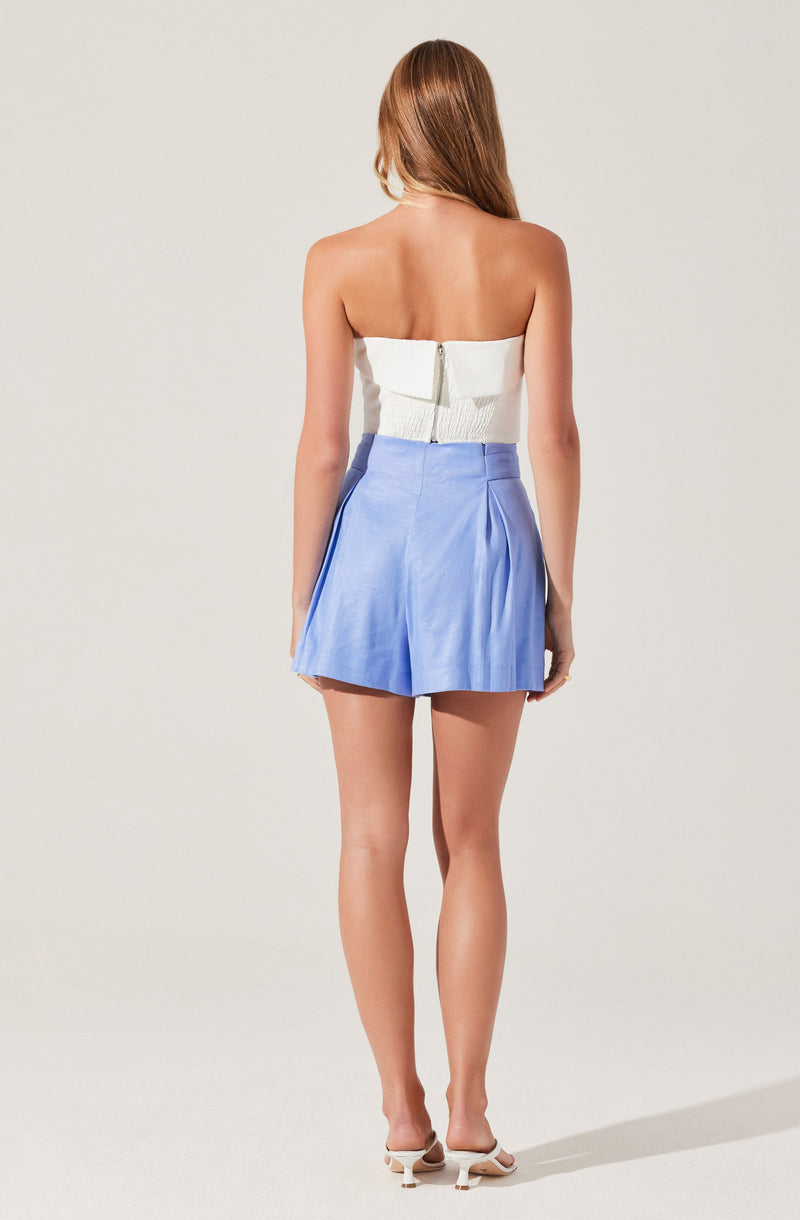A woman wearing a strapless white top with a back zipper and light blue high-waisted shorts. The shorts feature pleats on the front and a slightly flared silhouette. She is also wearing white heeled shoes.