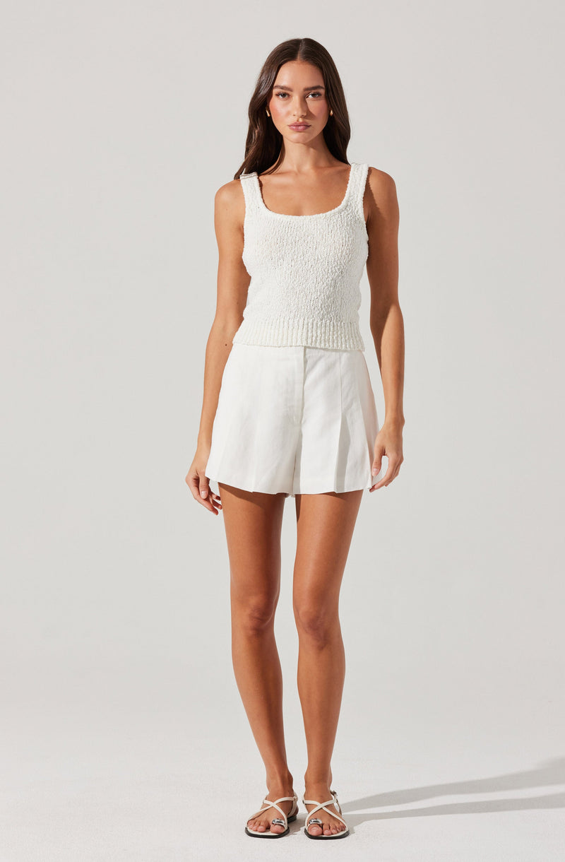 A woman is wearing a sleeveless, textured white knit top with a square neckline and wide shoulder straps. She is also dressed in high-waisted, white, pleated shorts. Her outfit is completed with white, strappy sandals.