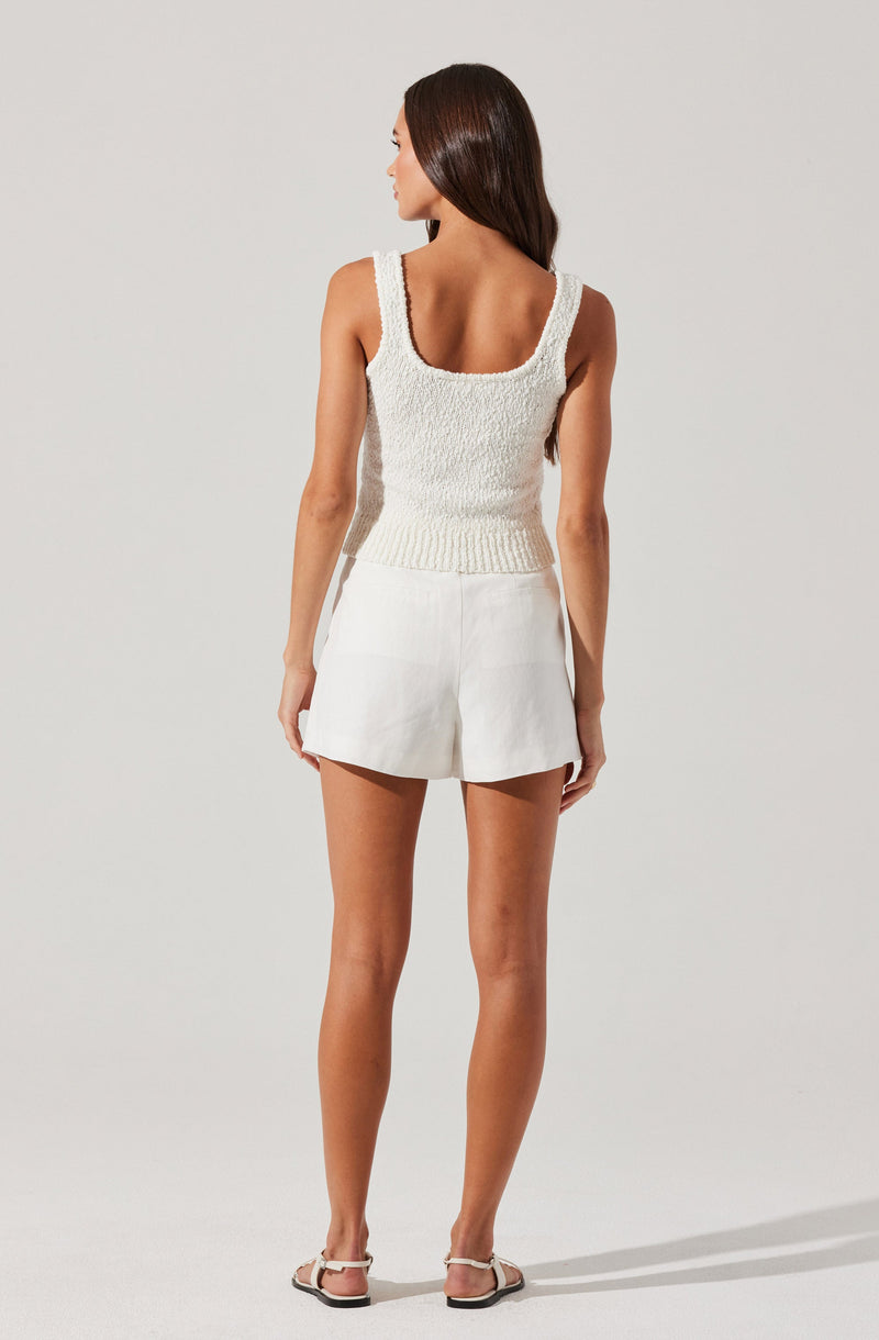 A woman is wearing a sleeveless, textured white tank top with wide straps and a fitted waist. She pairs it with high-waisted white shorts that have a relaxed fit. She completes the outfit with flat, open-toed sandals that have light-colored straps.