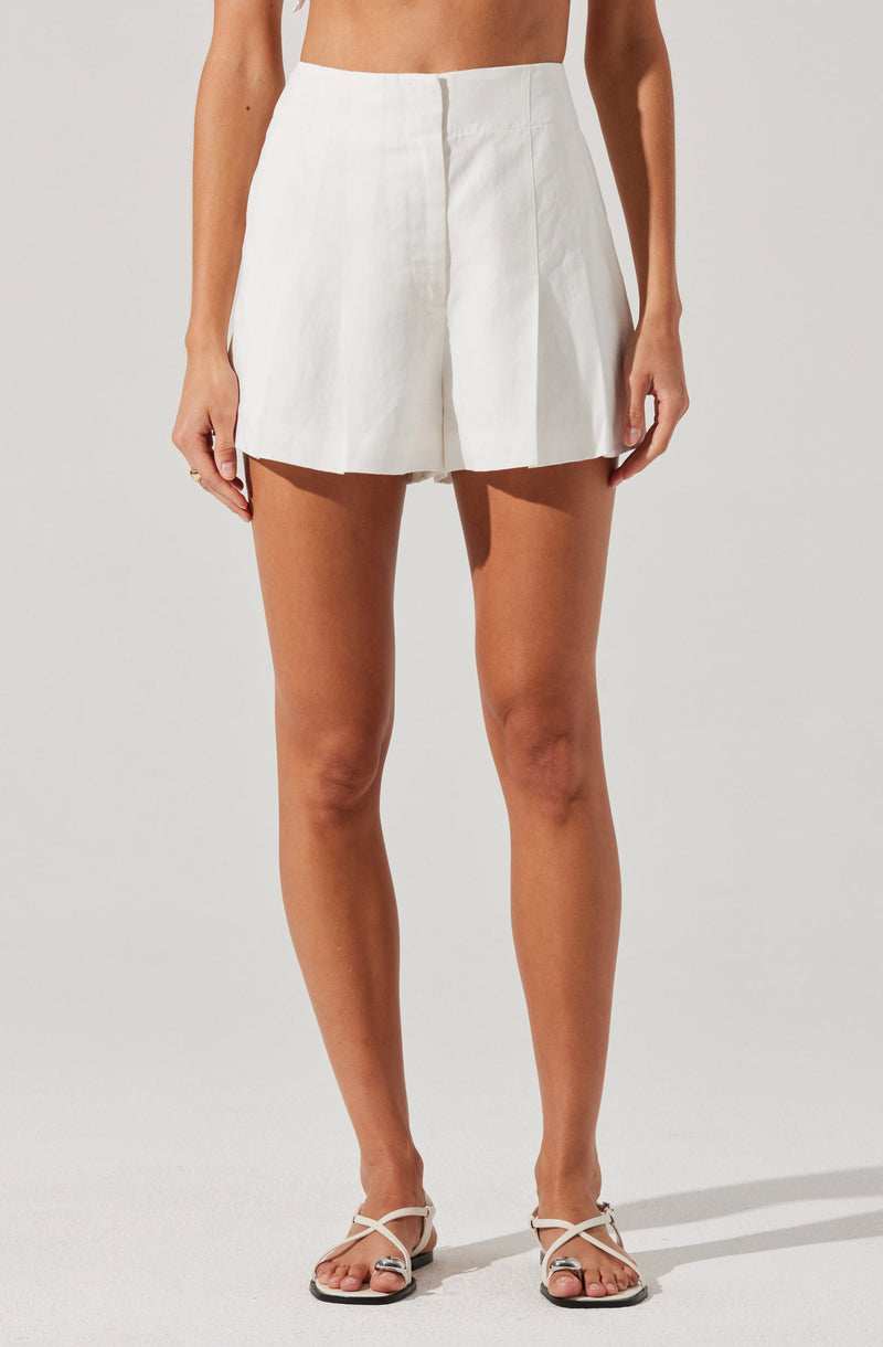 A woman is wearing high-waisted, white, pleated shorts made from a lightweight, linen-like fabric. The shorts have a front zipper closure with no visible pockets or embellishments. She is also wearing white strappy sandals with a small heel.