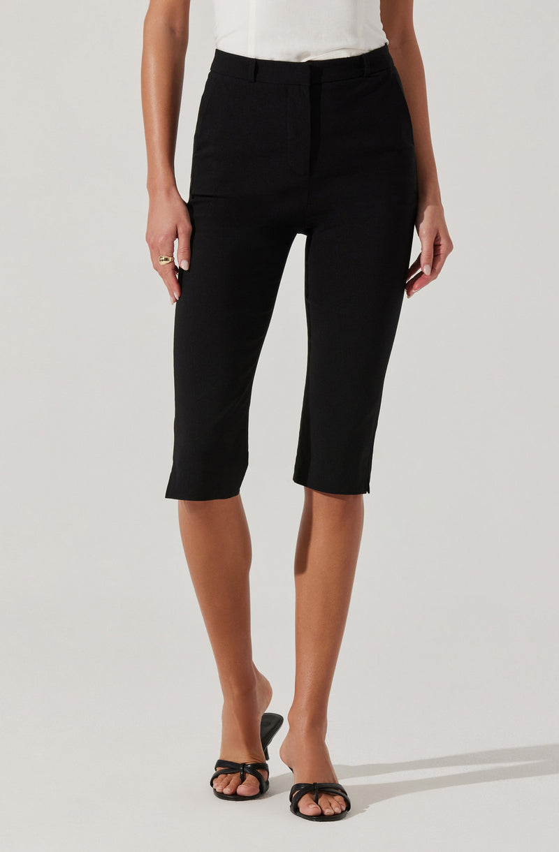 A woman is wearing black cropped dress pants with a high waist and a zipper front, paired with a white sleeveless top. She is also wearing black open-toe high-heeled sandals with thin straps.