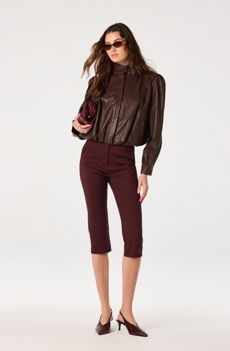 Woman in burgundy cropped pants, dark brown leather jacket, pointed heels, and sunglasses, holding a burgundy clutch.
