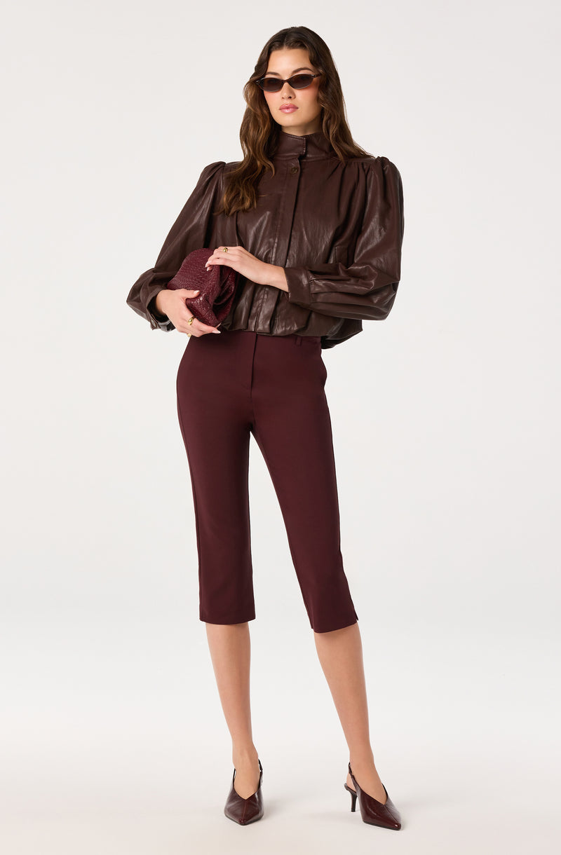 Woman in brown leather jacket, burgundy cropped pants, and dark heels, holding a clutch and wearing sunglasses.