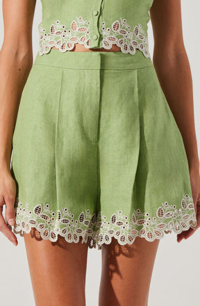 A woman is wearing a light green sleeveless top with decorative white floral embroidery along the hem and button area. The matching high-waisted shorts are also light green with the same white embroidered floral design along the hem, featuring intricate patterns of leaves and flowers.