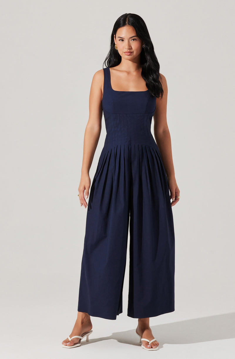 A woman is wearing a sleeveless navy blue jumpsuit with a square neckline. The fitted bodice features vertical seam details, and the wide-leg pants have pleats starting at the waist, creating a flowing silhouette. She is also wearing white heeled sandals.