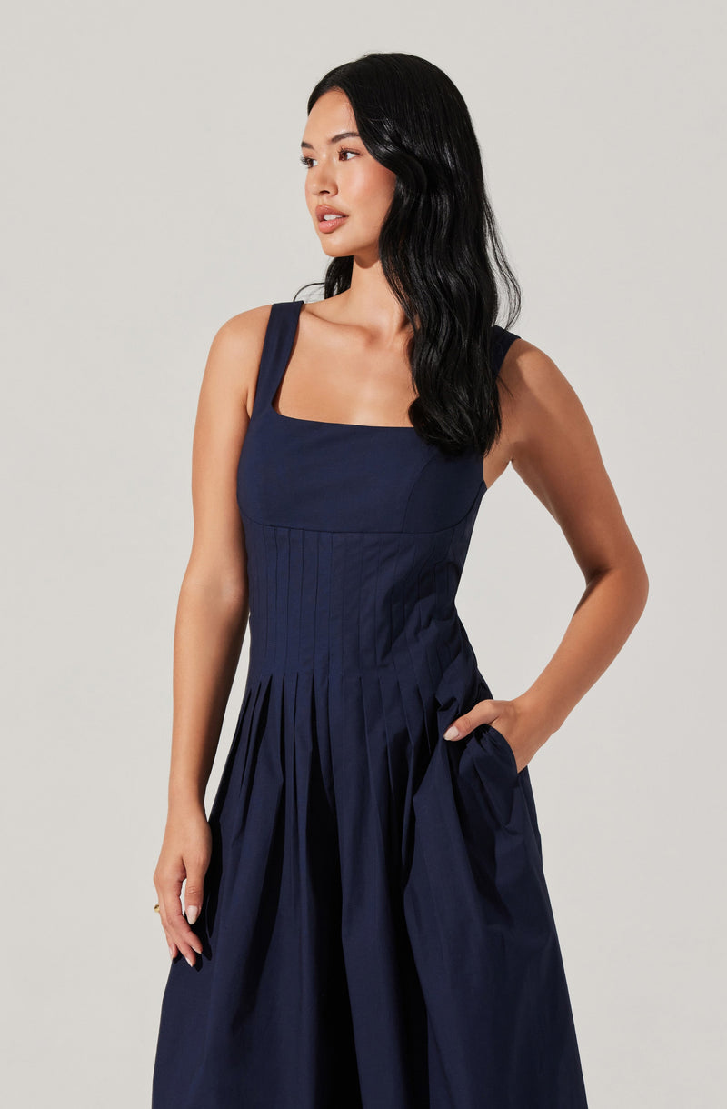 A woman is wearing a sleeveless, navy blue dress with a fitted bodice featuring wide straps and a square neckline. The dress has vertical pleats starting at the waist and extending down the skirt, which is full and gathered at the waist with some fabric in her pocket.