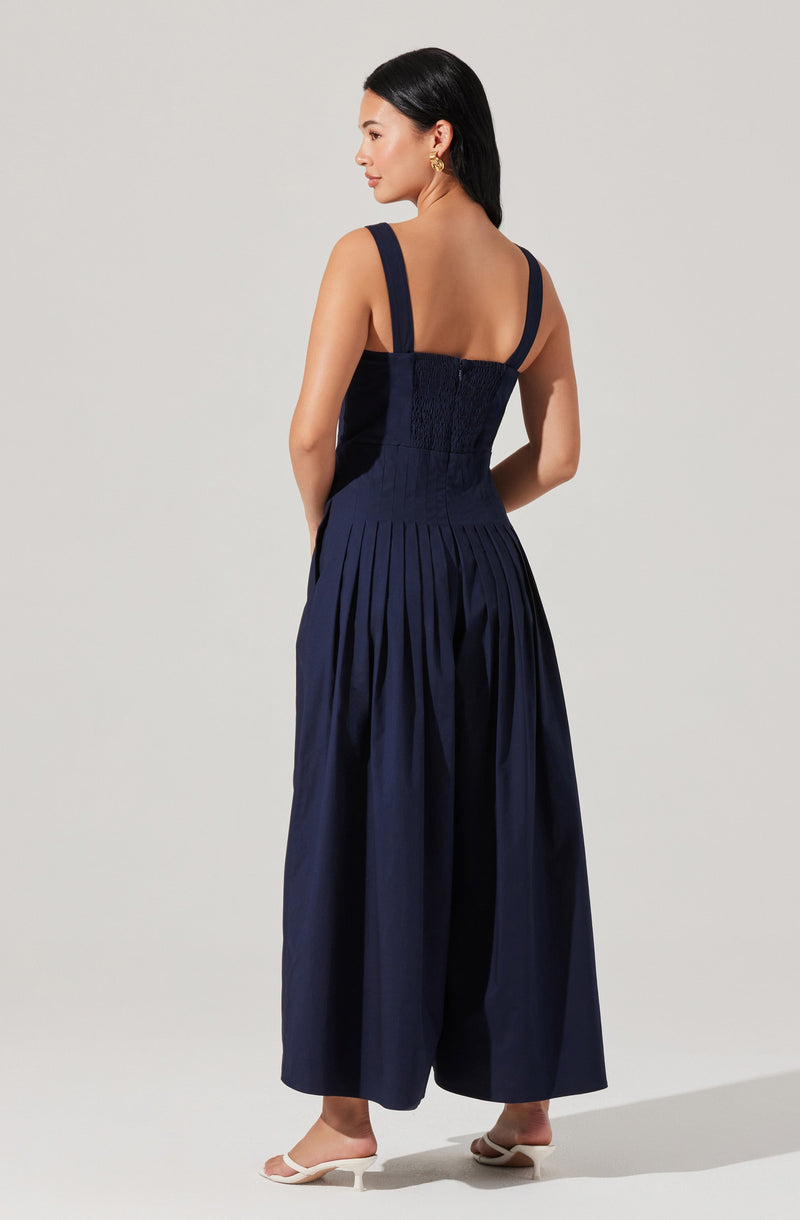A woman is wearing a dark navy blue sleeveless dress with wide shoulder straps. The bodice is fitted with smocking detail, and the skirt features wide pleats that add volume and structure. She pairs the outfit with light-colored open-toe heels.