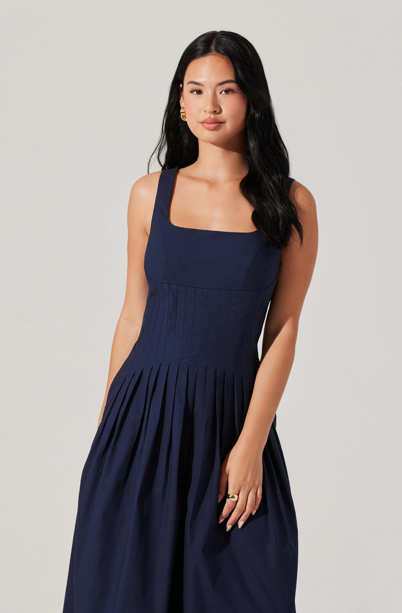 A woman is wearing a sleeveless navy blue dress with wide shoulder straps. The dress features a fitted bodice with subtle vertical seam details and a square neckline. The skirt has gathered pleats starting at the waist, creating a full, flowing silhouette. She has long, wavy black hair and is accessorized with gold hoop earrings and a gold ring.