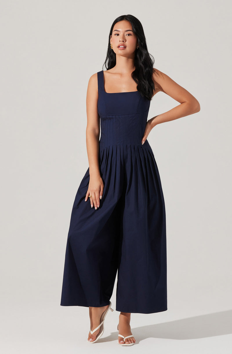 A woman is wearing a navy blue sleeveless jumpsuit with wide-leg pants. The jumpsuit features a fitted bodice with vertical pleats and wide shoulder straps. She is also wearing white high-heeled sandals.