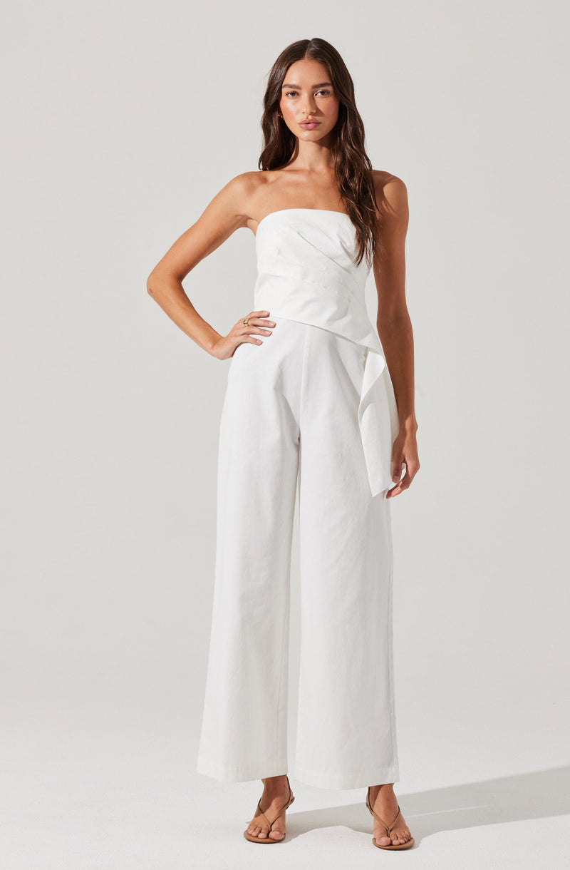 A woman is wearing a strapless white jumpsuit with wide-leg pants. The top is fitted with a slight drape, and there is a waist tie or peplum detail on the side. The overall look is elegant and minimalist. She is also wearing nude high-heeled sandals.