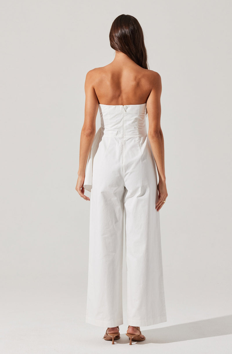 A woman is wearing a strapless white jumpsuit with a fitted bodice and wide-leg pants. The jumpsuit has a zipper closure at the back. She is also wearing nude high-heeled shoes.
