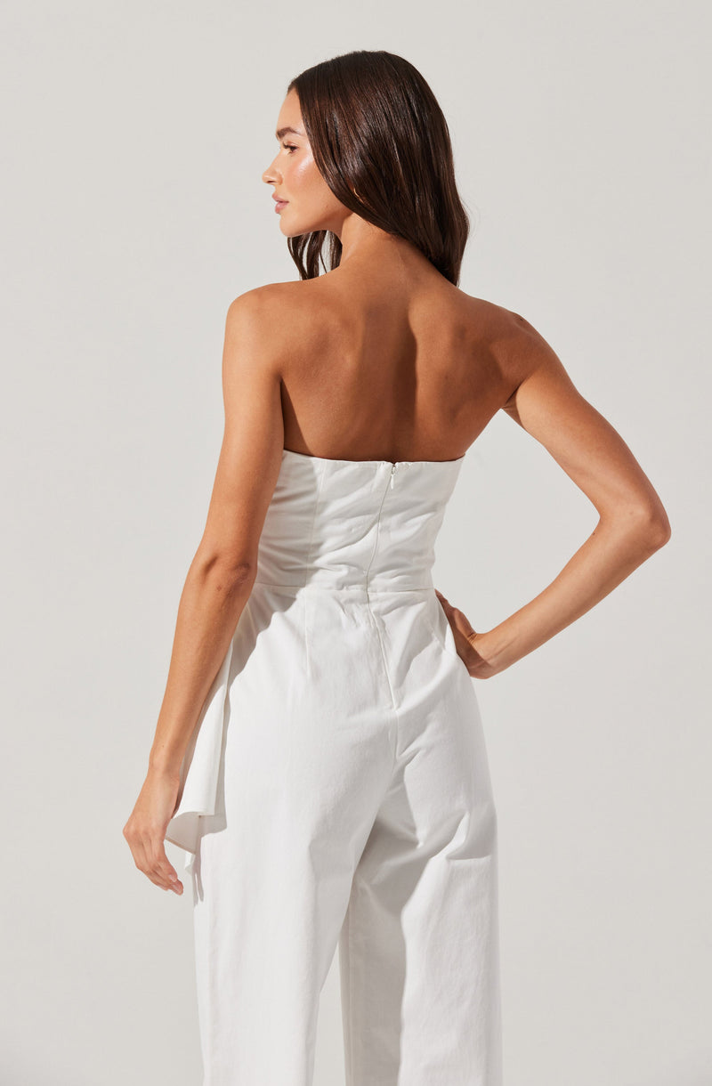 A woman is wearing a white, strapless jumpsuit with a fitted bodice and wide-leg pants. The jumpsuit features a hidden zipper at the back. She is posing with her left hand on her hip and her right arm hanging down, with her long dark hair styled straight.