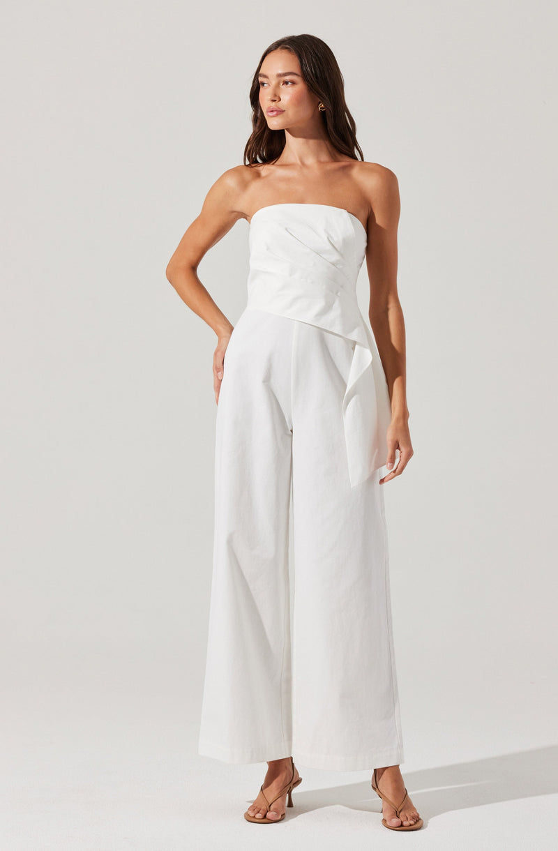 The woman is wearing a strapless, white jumpsuit with a fitted bodice that has a subtle pleated design. The jumpsuit features high-waisted, wide-leg pants that reach the floor and a decorative draped detail on one side of the waist. She is also wearing nude, open-toed high-heeled sandals.