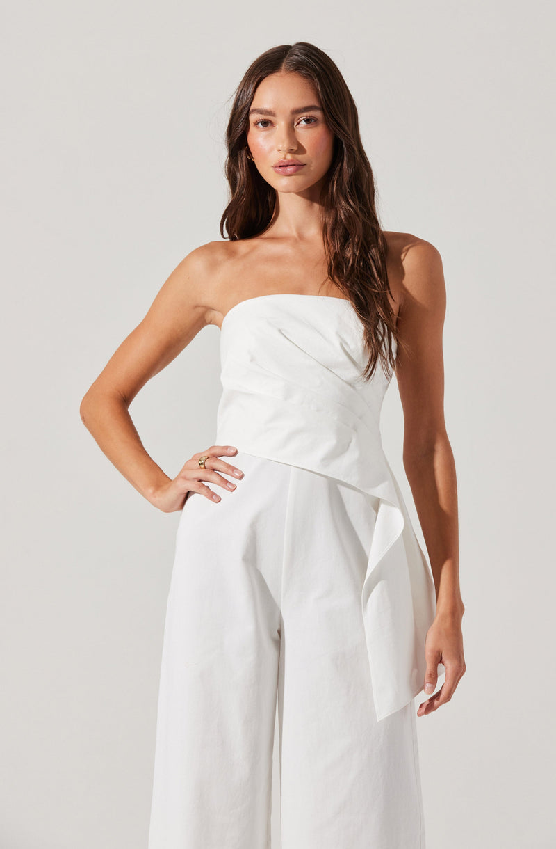 A woman is wearing a strapless white jumpsuit. The top is fitted and appears to be made of a slightly textured fabric with a subtle wrap design. The pants are wide-legged and high-waisted, made of a smooth, flowing fabric, with a large, decorative bow or sash tied on the right side.