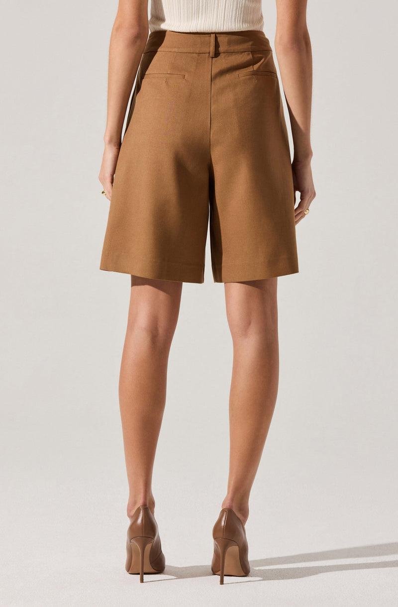 The image shows a person wearing high-waisted, knee-length brown shorts with two back pockets. They are paired with a sleeveless ribbed top in a light neutral color and beige high-heeled shoes.