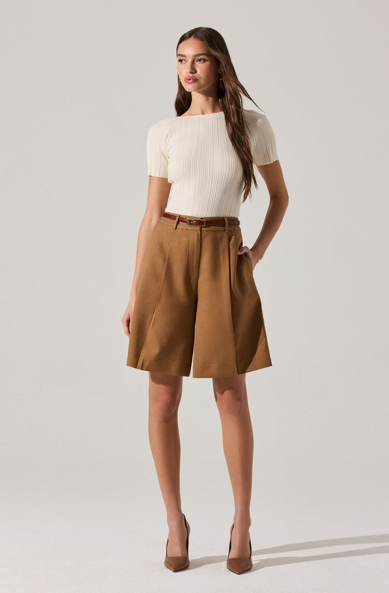 A woman is wearing a cream-colored, ribbed short-sleeve top with a boat neckline, tucked into high-waisted, tan knee-length shorts with pleats on the front. She is accessorized with a thin brown belt and is wearing nude-colored pointed-toe high heels.