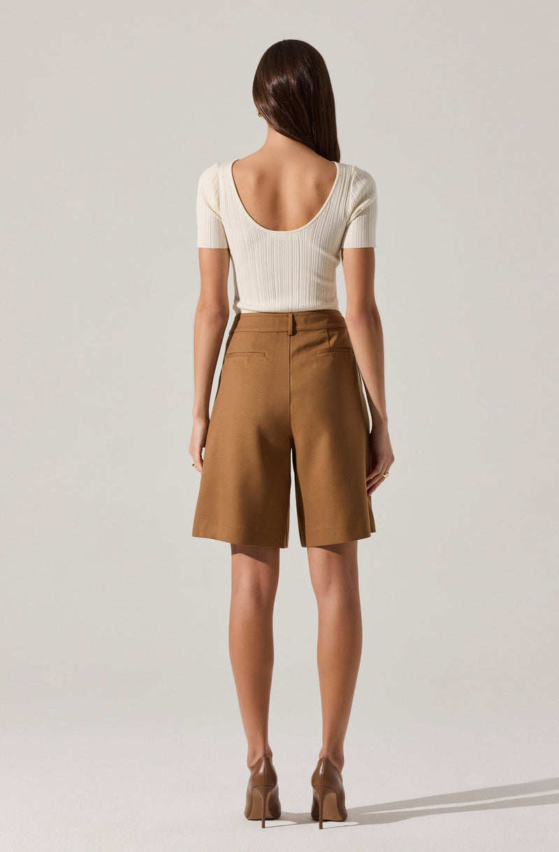 A woman is wearing a cream-colored, short-sleeved, ribbed knit top with a wide scoop neckline. She pairs it with high-waisted, knee-length, camel-colored shorts that feature a smooth waistband and side seam pockets. She completes the outfit with nude high-heeled shoes.