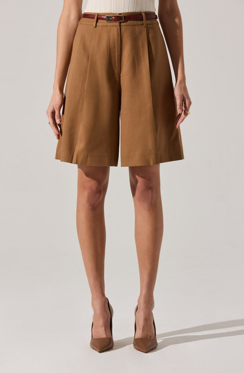 A person is wearing high-waisted, wide-leg knee-length brown shorts with pleats. They have a slim belt with a gold buckle. The individual is also wearing nude-colored high heels. Their left hand has a gold ring on the ring finger.