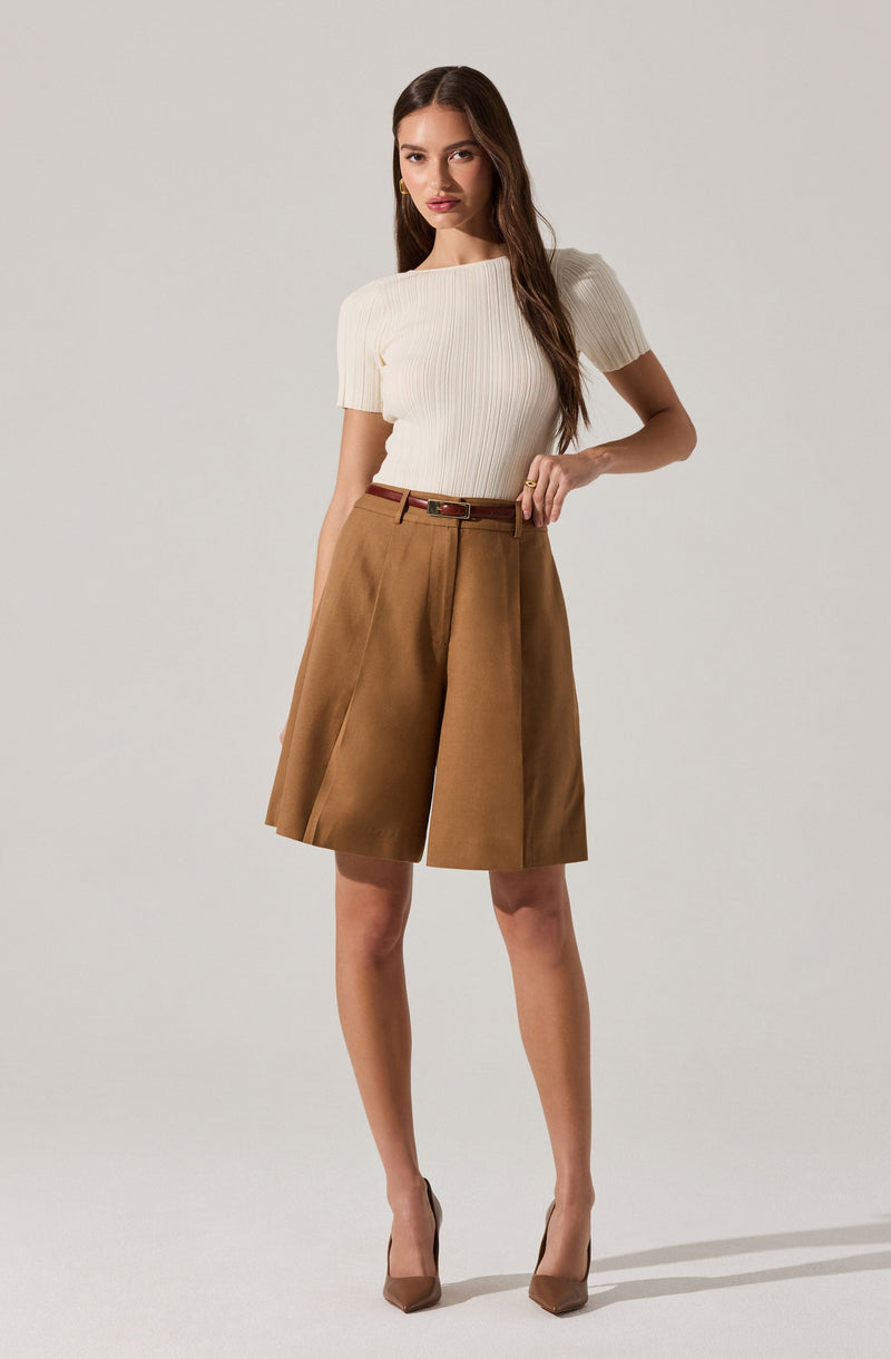 A woman is wearing a short-sleeved, ribbed, cream-colored top with a boat neckline. She pairs it with high-waisted, wide-legged, camel-colored culottes that have front pleats and are secured with a belt. She completes the outfit with taupe pointed-toe high heels.