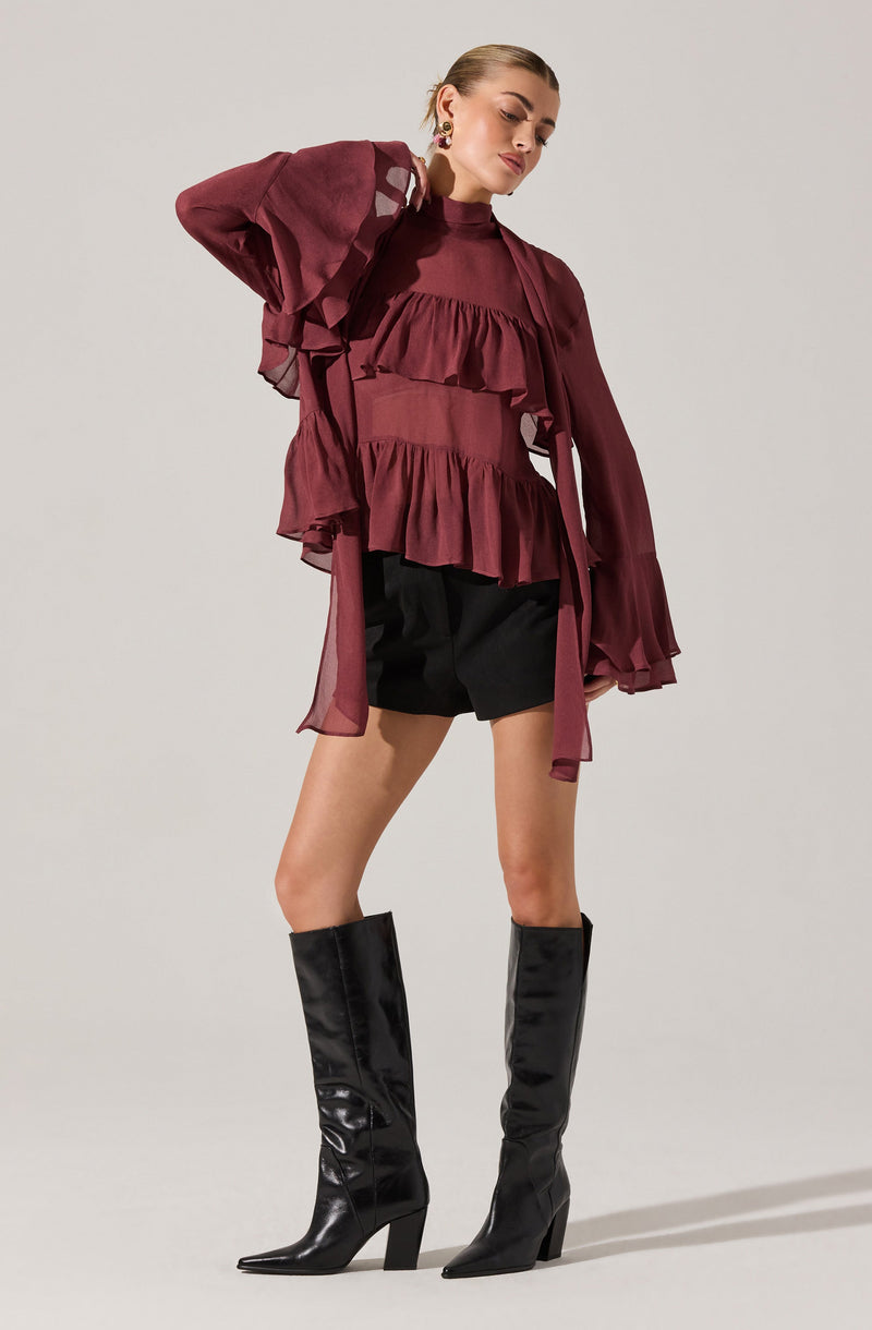 A woman is wearing a long-sleeved, high-neck, burgundy blouse with ruffled layers across the chest and front. The sleeves are sheer and billowy, gathered at the cuffs. She pairs the blouse with black shorts and tall, shiny black leather boots with high heels.