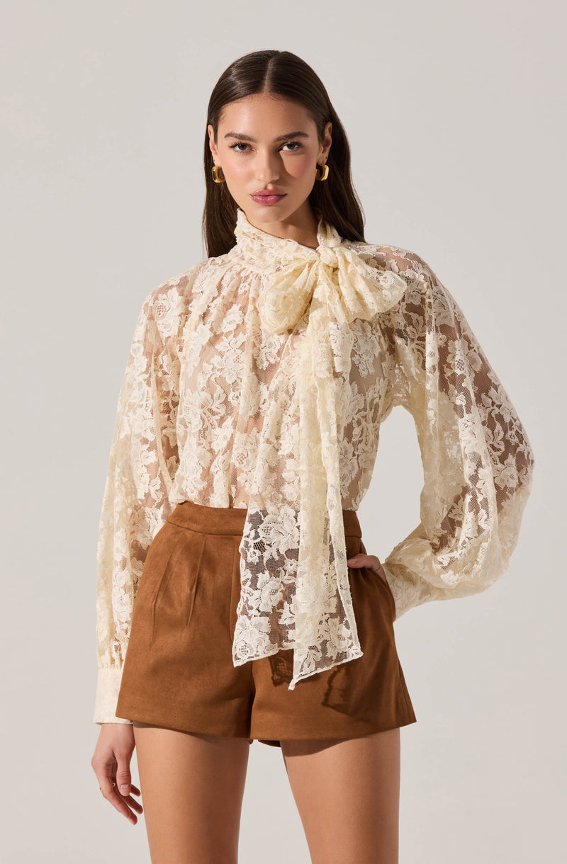 Woman wearing a cream lace blouse with a bow tie collar and brown shorts.