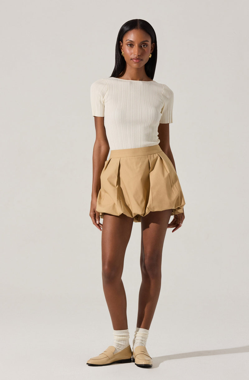 A woman is wearing a fitted, short-sleeved, ribbed, cream-colored top with slightly puffed sleeves. She pairs it with high-waisted, beige, pleated shorts that are loose-fitting and have a unique, rounded, and gathered design. She completes her outfit with light-colored socks and beige slip-on shoes.