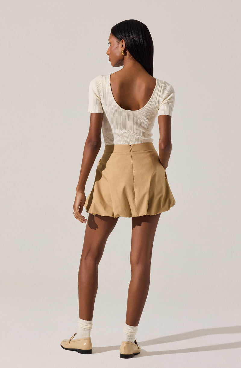 A woman is wearing a fitted, cream-colored, ribbed knit top with a low, rounded back and short sleeves. She pairs it with a beige, high-waisted, pleated mini skirt with a zipper at the back. She completes her outfit with cream-colored ankle socks and matching beige shoes. She has gold earrings and has long, straight black hair.