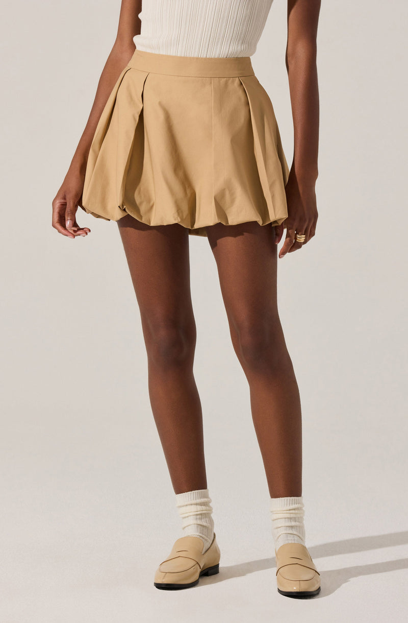 A person is wearing a high-waisted beige pleated skirt with large, gathered pleats creating a balloon effect. They are also wearing a sleeveless, ribbed, beige top tucked into the skirt. The outfit is complemented with beige loafers and cream-colored socks, and the person has a ring on their right hand.