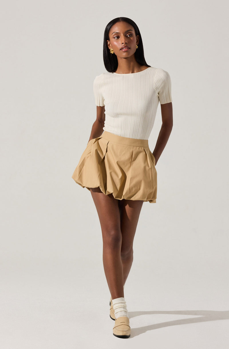 A woman is wearing a fitted, short-sleeved, ribbed cream-colored top with a round neckline. She has on a pair of high-waisted, tan-colored shorts with a voluminous, gathered, and pleated design that creates a bubble shape. She is also wearing beige ankle socks and beige shoes.