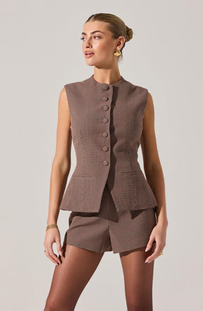 A woman is wearing a sleeveless, tailored vest and matching shorts made of fabric with a small checkered pattern in brown, black, and white. The vest has a high neckline with a row of fabric-covered buttons down the front, and the shorts have a tailored fit with a subtle pleat detail.