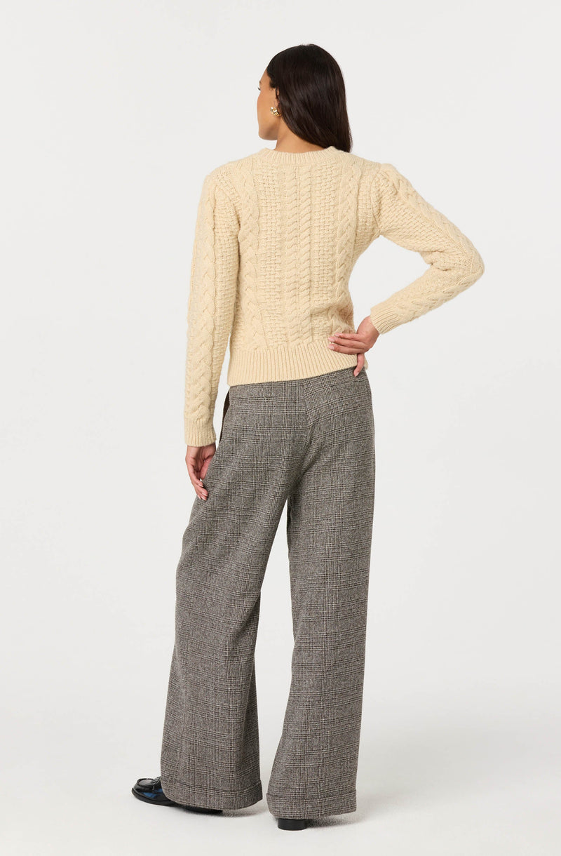 A woman is wearing a cream-colored, chunky cable-knit sweater with long sleeves and a ribbed hem. She is also dressed in high-waisted, wide-leg plaid trousers in shades of brown and gray, paired with black shoes.