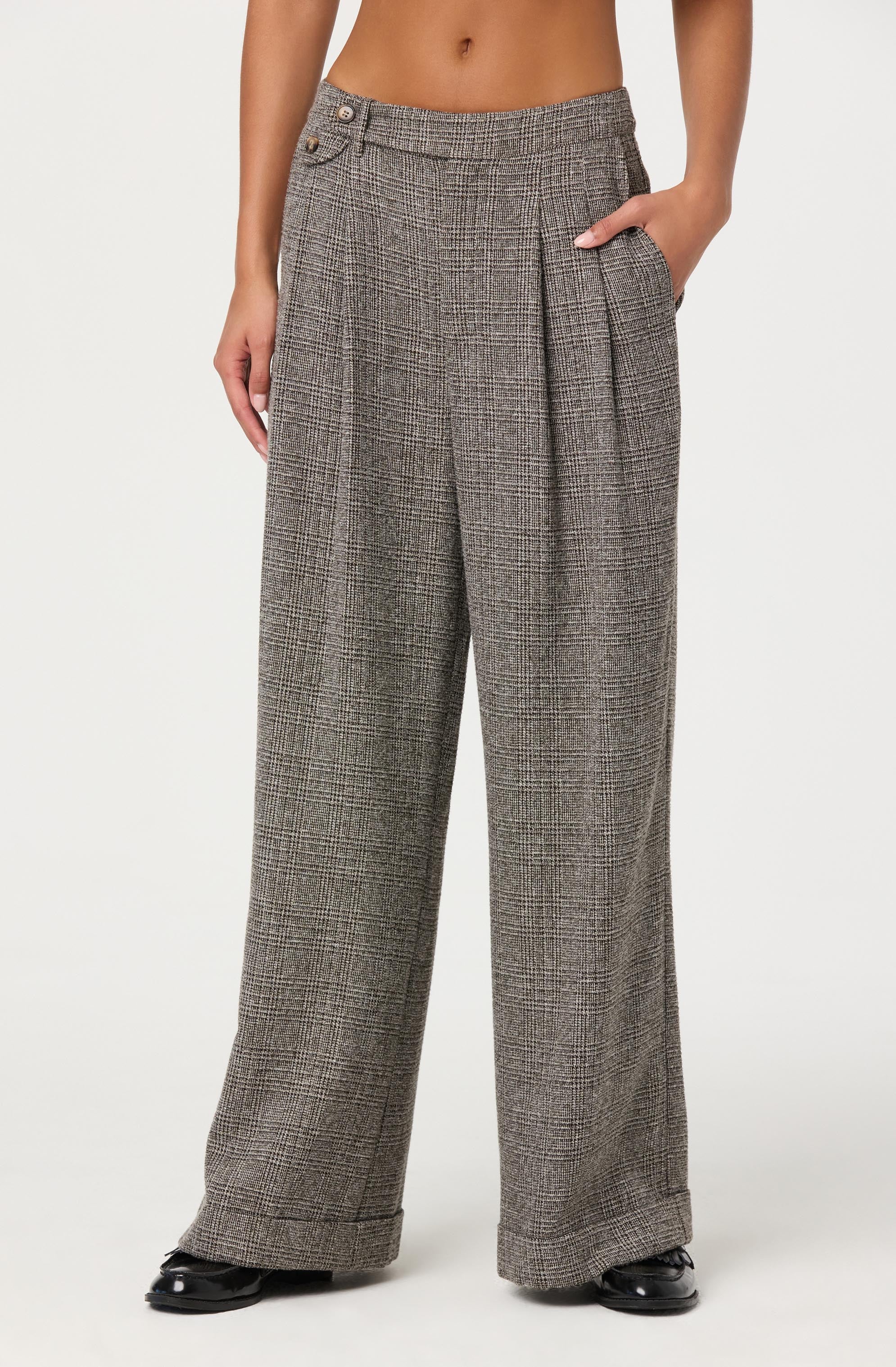 A person is wearing high-waisted, wide-leg plaid pants in shades of brown, gray, and black. The pants feature pleats at the waist, a side button closure near the waistband, and a front pocket on the right side. The person has one hand in the pocket and is standing with their legs slightly apart. They are also wearing black shoes.