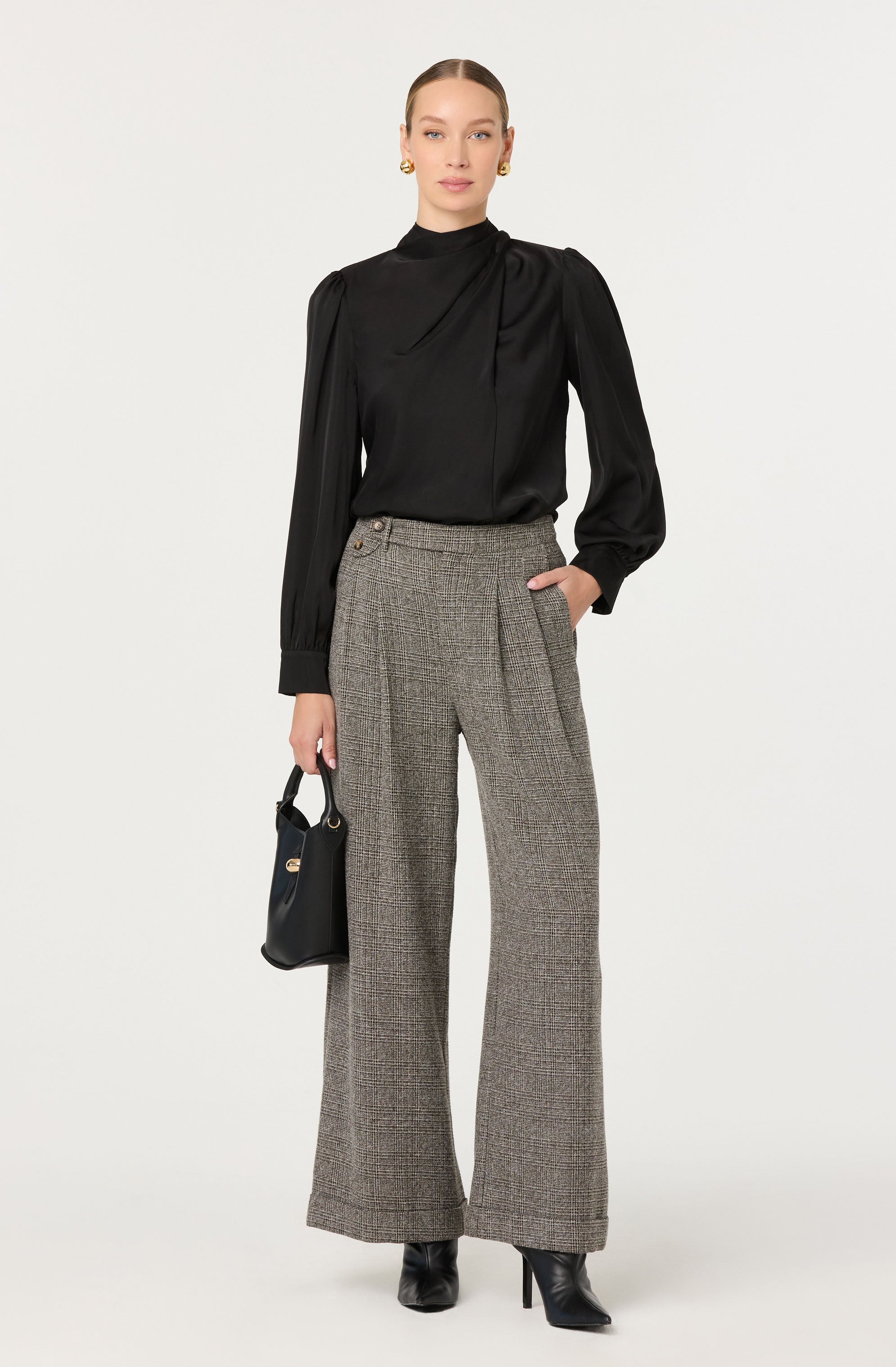 The woman is wearing a black long-sleeved blouse with a high neckline and subtle pleats at the shoulder, paired with high-waisted, wide-leg plaid trousers in shades of gray and brown. She accessorizes with gold hoop earrings and holds a black handbag with gold hardware. She completes the look with black pointed-toe high-heeled boots.