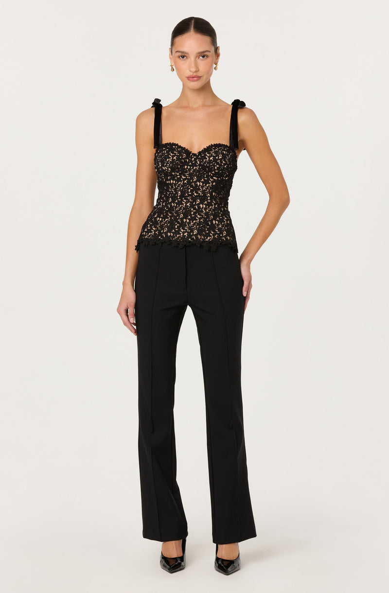 A woman is wearing a sleeveless black top with lace details and shoulder straps tied into bows. She pairs it with high-waisted black trousers that have a straight-leg fit. She completes the look with black pointed heels.