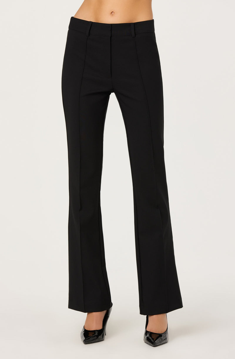 A person is wearing black dress pants with a high waist and belt loops. The pants have a tailored fit with a crease running down the front of each leg and are slightly flared at the hem. The person is also wearing black high-heeled shoes.