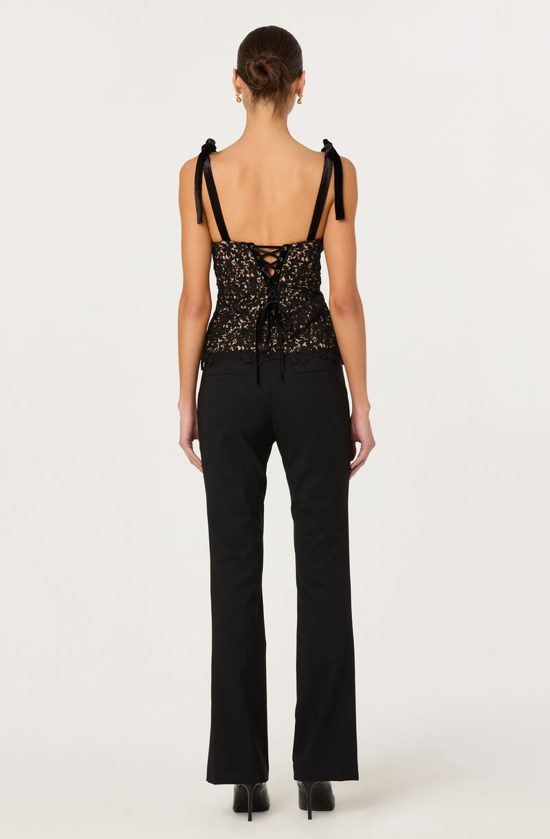 The woman is wearing a sleeveless, black lace top with shoulder ties and a lace-up back detail. She pairs this with black, high-waisted, straight-leg pants and black high-heeled shoes.
