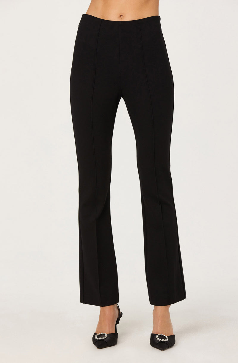 A pair of black dress pants with a high waist and tailored fit. The pants have a wide leg design that falls straight to the hem, creating a sleek and polished appearance. The fabric appears smooth and structured. The model is wearing the pants with black shoes that have decorative embellishments on the toes.