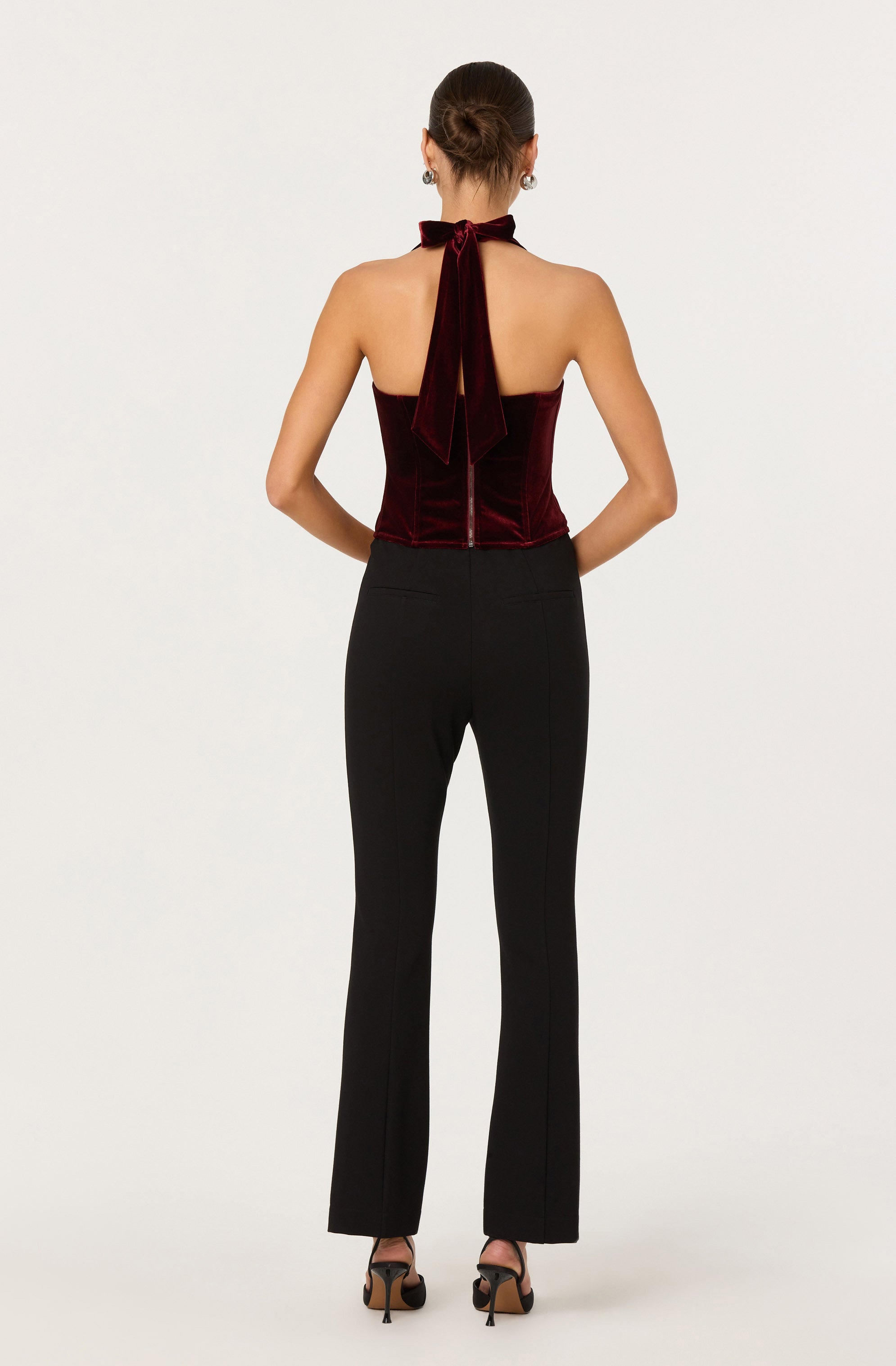 The image shows a woman dressed in a sleeveless, deep red velvet top with a halter neck and a bow tie at the neck. She is also wearing high-waisted, black tailored pants that are fitted through the hips and thighs with a slight flare at the bottom. She is wearing black high-heeled sandals.