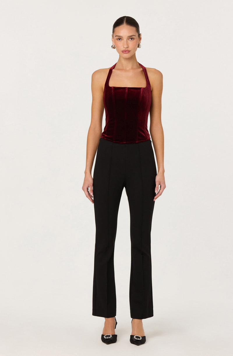 The woman is wearing a fitted, sleeveless, burgundy velvet top with a square neckline and halter straps. She pairs it with high-waisted black dress pants that have a tailored fit and slight flare at the hem. She is also wearing black pointed-toe heels with a decorative embellishment on the front.