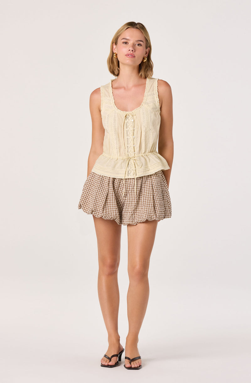 Woman wearing a cream lace tank top and brown gingham shorts.