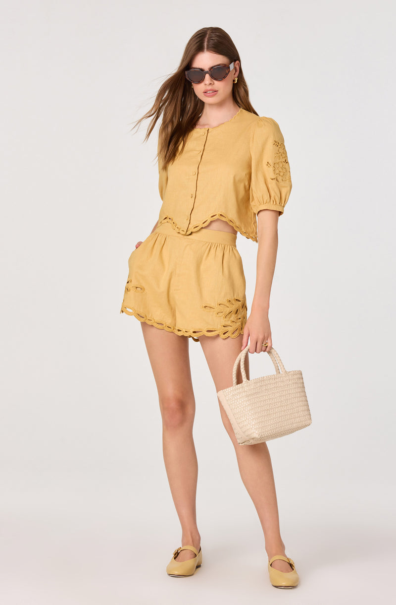 Woman in yellow cutout top and shorts, carrying a woven bag and wearing sunglasses.