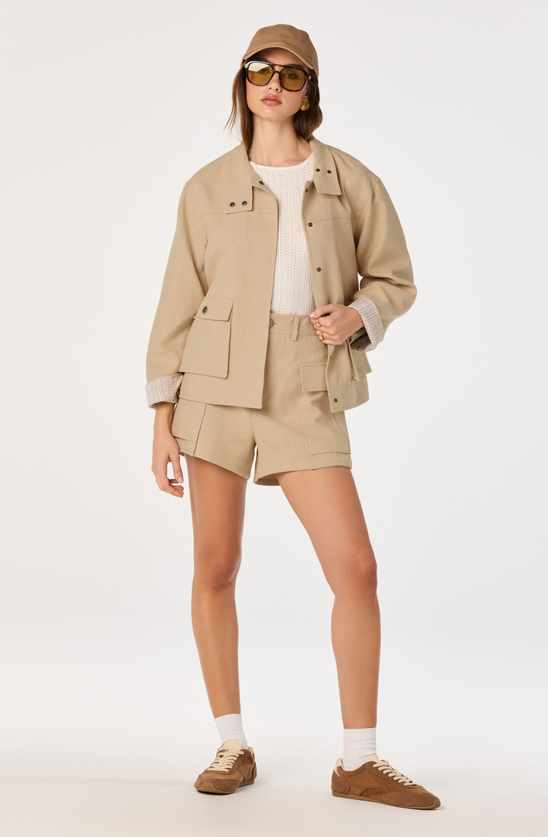 Woman wearing beige jacket, matching shorts, white sweater, brown cap, sunglasses, and sneakers.