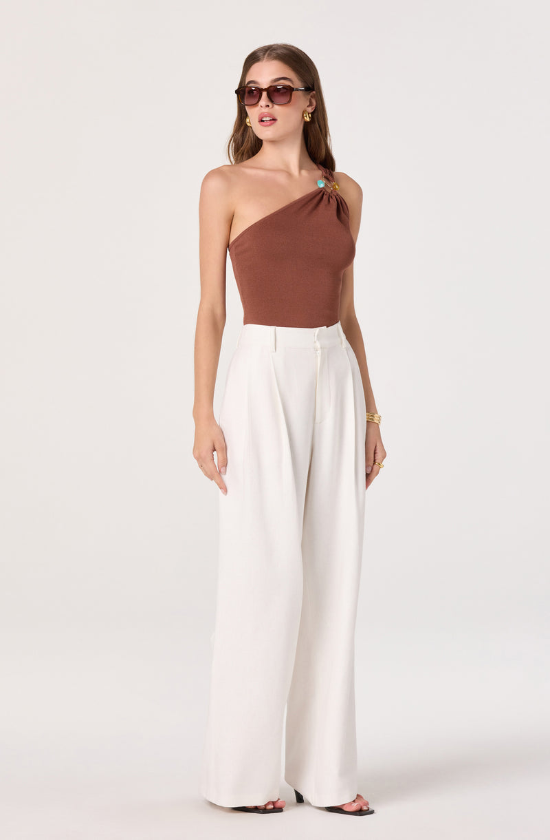 Brown one-shoulder top with decorative shoulder detail paired with high-waisted white wide-leg trousers.