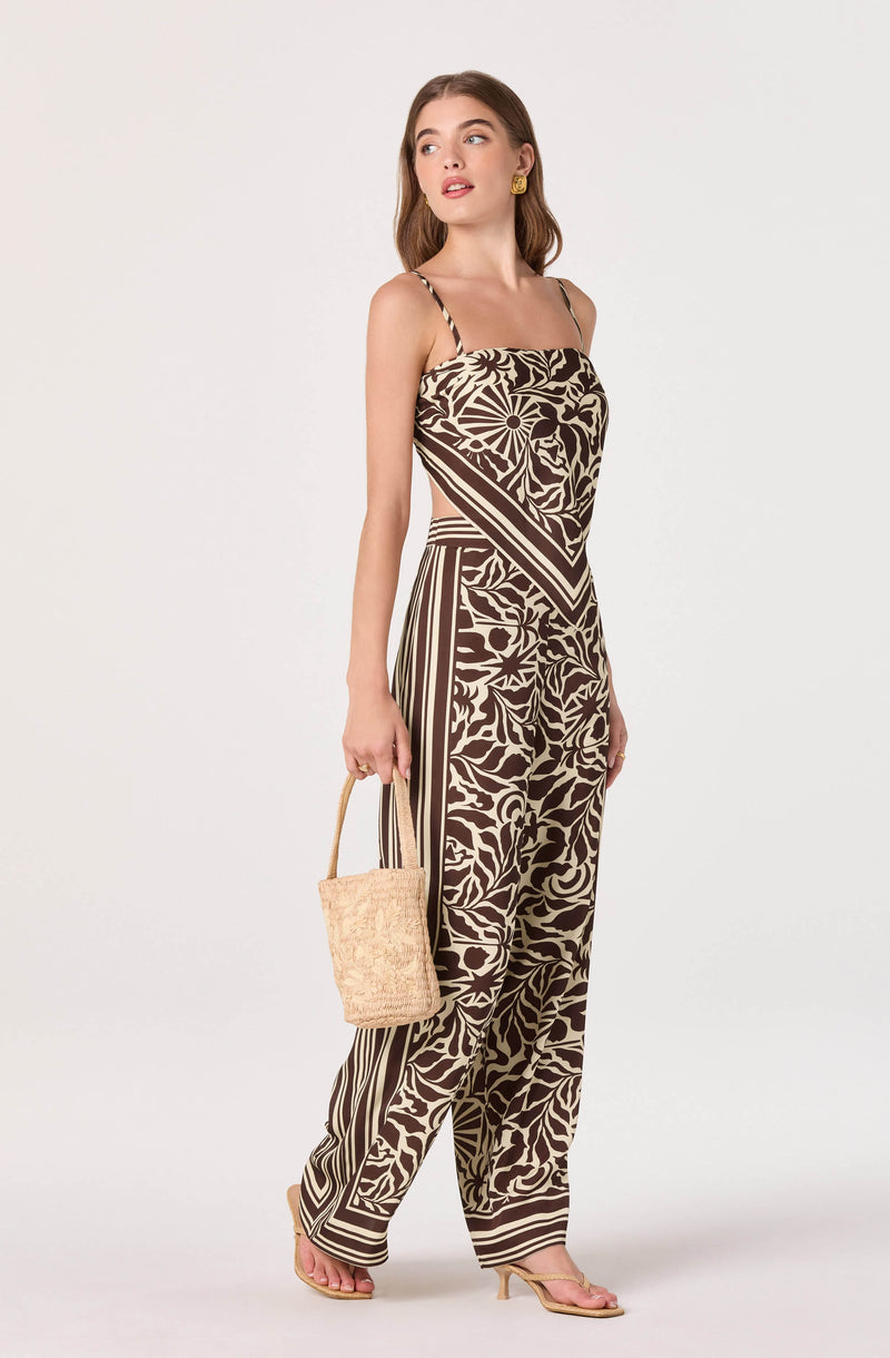 Woman wearing a beige and brown patterned sleeveless jumpsuit with thin straps, holding a beige textured handbag.