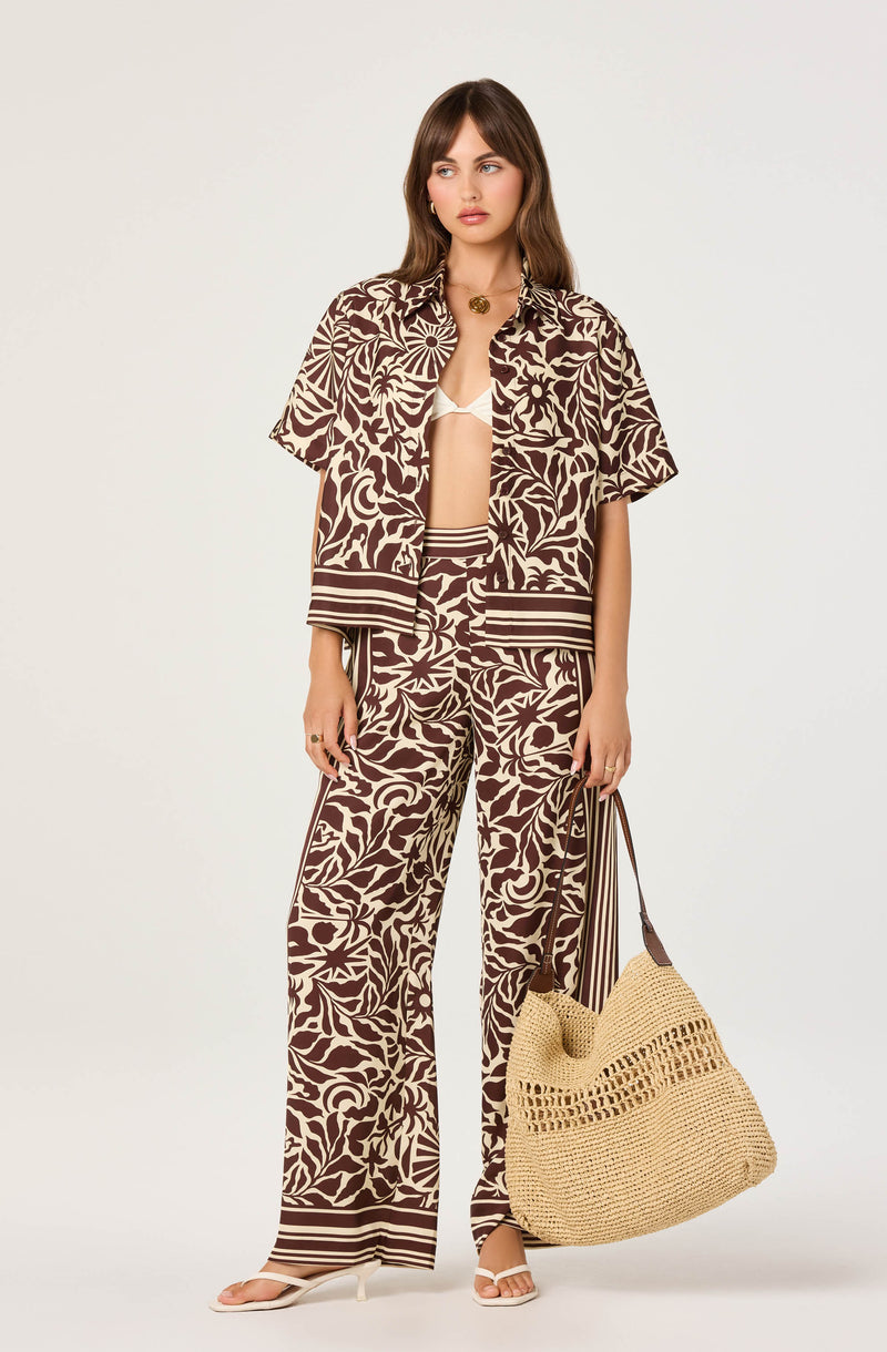 Woman wearing a brown and beige leaf-patterned shirt and wide-leg pants, holding a woven bag.