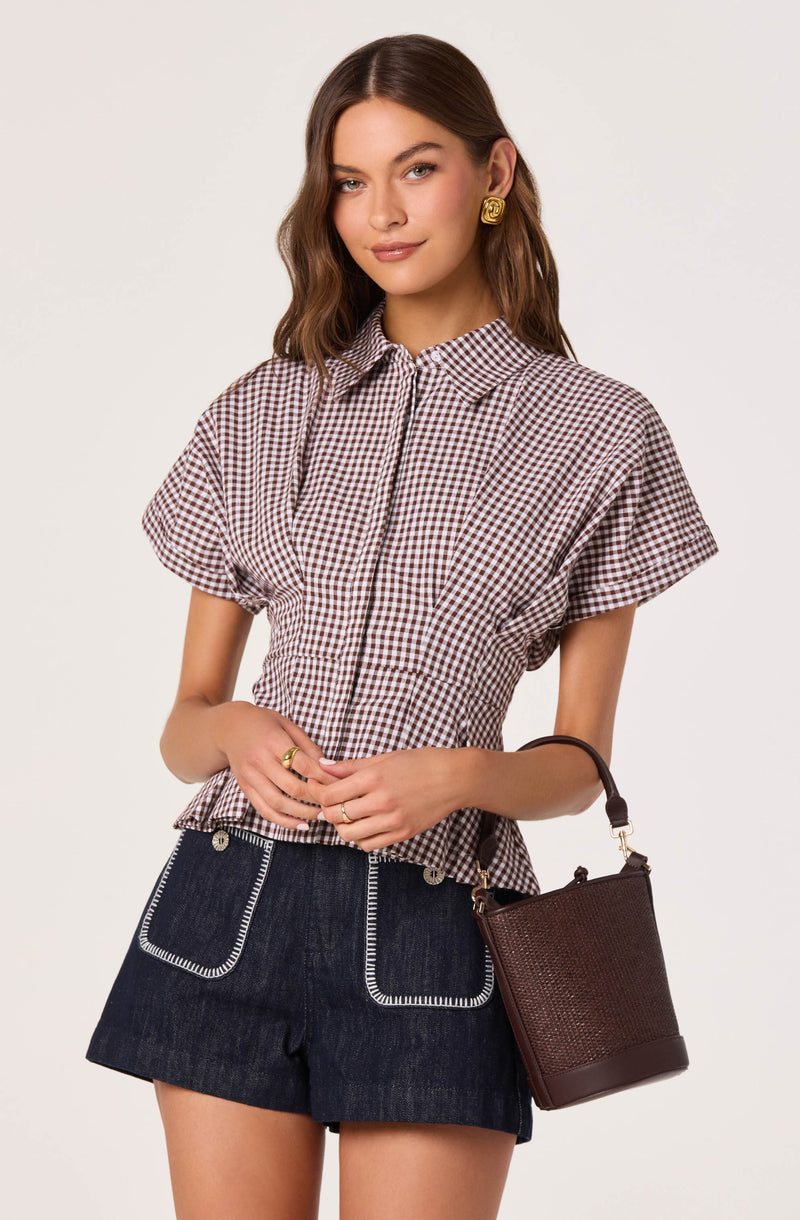 Woman wearing a brown checkered short-sleeve shirt with collar and dark denim shorts with white stitch details.