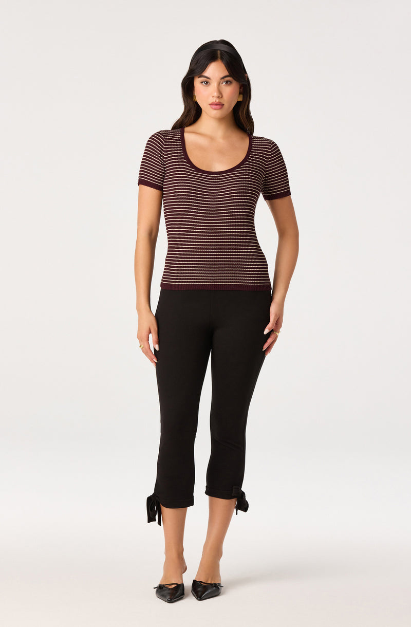 Woman wearing a short-sleeve, striped maroon and beige top with black cropped pants with bow details at the hem.